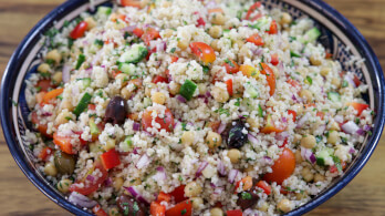 A colorful bowl of couscous salad filled with diced tomatoes, cucumbers, red onions, bell peppers, chickpeas, black olives, and herbs. The ingredients are mixed together, creating a vibrant and fresh-looking dish.