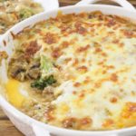 A white baking dish filled with a cheesy chicken and broccoli casserole, with a portion scooped out and placed on a white plate in the background. The casserole has a golden-brown, bubbly cheese topping. The dish sits on a wooden table.