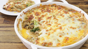 A white baking dish filled with a cheesy chicken and broccoli casserole, with a portion scooped out and placed on a white plate in the background. The casserole has a golden-brown, bubbly cheese topping. The dish sits on a wooden table.