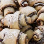 A plate of rugelach pastries arranged closely together on a decorative plate. The pastries are rolled with layered filling, dusted with powdered sugar, and have a golden-brown crust. The background shows a wooden surface.