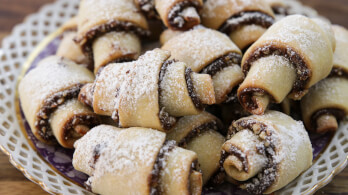 A plate of rugelach pastries arranged closely together on a decorative plate. The pastries are rolled with layered filling, dusted with powdered sugar, and have a golden-brown crust. The background shows a wooden surface.