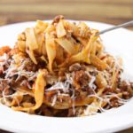 A close-up of a white plate filled with fettuccine pasta topped with meat sauce and sprinkled with grated cheese. A hand is lifting a forkful of pasta from the plate. The background features a wooden table.