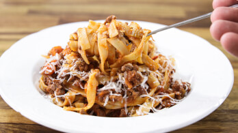 A close-up of a white plate filled with fettuccine pasta topped with meat sauce and sprinkled with grated cheese. A hand is lifting a forkful of pasta from the plate. The background features a wooden table.