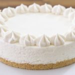 A creamy, round cheesecake with a graham cracker crust, decorated with dollops of whipped cream around the edge, sits on a white cake stand against a blurred wooden background.