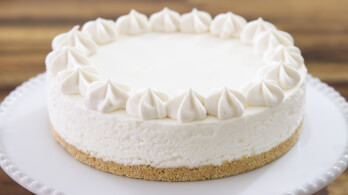 A creamy, round cheesecake with a graham cracker crust, decorated with dollops of whipped cream around the edge, sits on a white cake stand against a blurred wooden background.