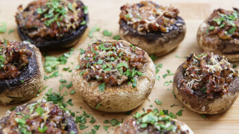 Close-up of stuffed mushrooms garnished with chopped fresh parsley, placed on a wooden board. The stuffing appears to be a savory mixture, possibly including ground meat and cheese, giving the mushrooms a slightly golden, crispy top.