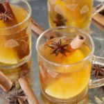 Three clear glass mugs filled with a spiced apple cider or similar beverage, each garnished with a star anise and a cinnamon stick. The mugs are on a reflective silver tray, accompanied by more star anise, cinnamon sticks, and a whole lemon.