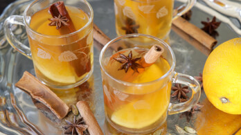 Three clear glass mugs filled with a spiced apple cider or similar beverage, each garnished with a star anise and a cinnamon stick. The mugs are on a reflective silver tray, accompanied by more star anise, cinnamon sticks, and a whole lemon.