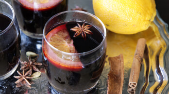 A close-up of glasses filled with mulled wine, garnished with slices of lemon and star anise. Next to the glasses are a whole lemon, cinnamon sticks, and additional star anise on top of a reflective surface.