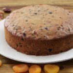 A round fruitcake sits on a white plate atop a wooden surface. Surrounding the plate are various dried fruits and nuts, including dried apricots, raisins, walnuts, and pecans. The cake shows bits of fruits and nuts embedded in its golden brown crust.