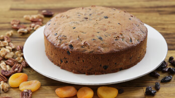 A round fruitcake sits on a white plate atop a wooden surface. Surrounding the plate are various dried fruits and nuts, including dried apricots, raisins, walnuts, and pecans. The cake shows bits of fruits and nuts embedded in its golden brown crust.