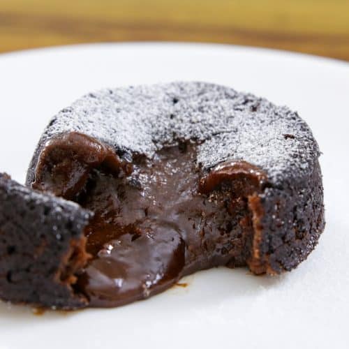 A close-up of a chocolate lava cake on a white plate. The cake is dusted with powdered sugar, and its molten chocolate center is oozing out, showing a rich and gooey texture. The background is slightly blurred, focusing attention on the dessert.