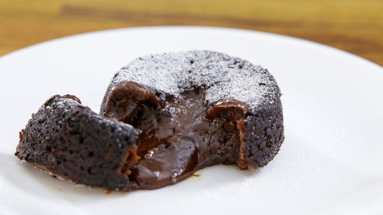 A close-up of a chocolate lava cake on a white plate. The cake is dusted with powdered sugar, and its molten chocolate center is oozing out, showing a rich and gooey texture. The background is slightly blurred, focusing attention on the dessert.