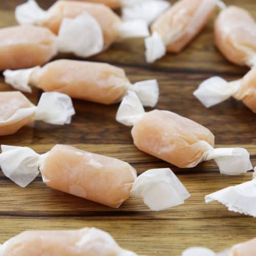 Several pieces of caramel-colored taffy are individually wrapped in white paper, scattered randomly on a wooden surface. The texture of the taffy appears soft, and the wrappers are twisted at both ends.