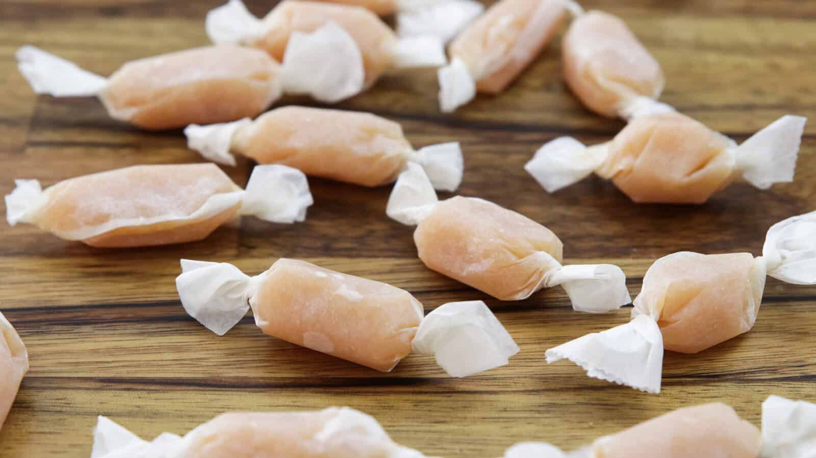 Several pieces of caramel-colored taffy are individually wrapped in white paper, scattered randomly on a wooden surface. The texture of the taffy appears soft, and the wrappers are twisted at both ends.