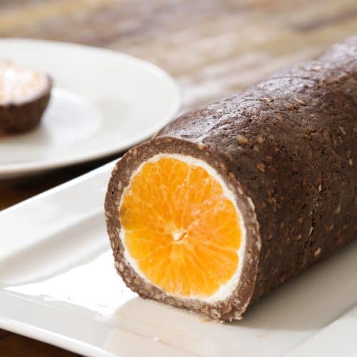 A sandwich-like dessert featuring a whole orange wrapped in a chocolate and nut mixture, sliced to expose the contrasting orange and brown layers, presented on a white rectangular plate. Another slice of the dessert is placed on a separate white plate in the background.