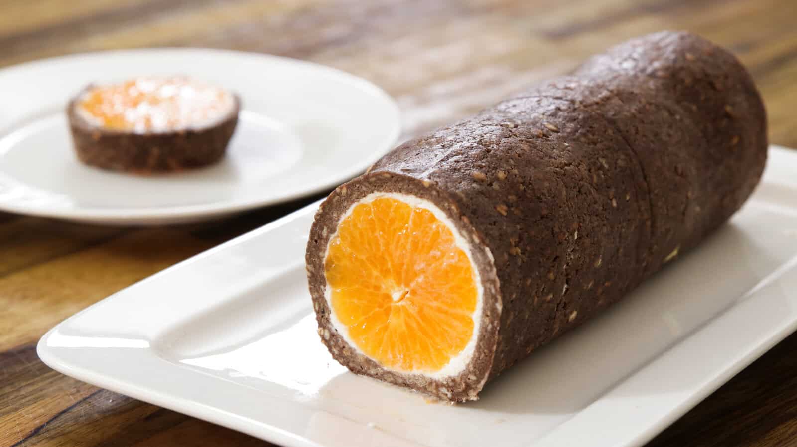 A sandwich-like dessert featuring a whole orange wrapped in a chocolate and nut mixture, sliced to expose the contrasting orange and brown layers, presented on a white rectangular plate. Another slice of the dessert is placed on a separate white plate in the background.