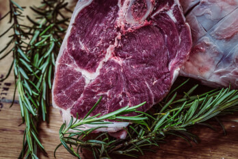 A raw steak on a wooden surface, garnished with sprigs of fresh rosemary. One sprig lies beside the steak while another is partially covering it.