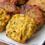 A close-up of a plate of golden-brown vegetable fritters. The fritters are slightly crispy on the outside, with visible flecks of green herbs and orange carrot pieces. The plate is set on a wooden surface with a small bowl of dipping sauce partially visible.