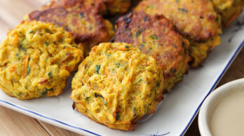 A close-up of a plate of golden-brown vegetable fritters. The fritters are slightly crispy on the outside, with visible flecks of green herbs and orange carrot pieces. The plate is set on a wooden surface with a small bowl of dipping sauce partially visible.