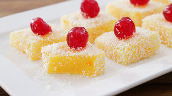 Squares of yellow cake topped with desiccated coconut and red glace cherries are arranged on a white rectangular plate. The yellow cakes have a smooth texture and are visually contrasted by the white coconut and bright red cherries.