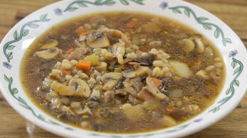 A bowl of mushroom barley soup with carrots, potatoes, and herbs, served in a white bowl with a green floral rim, placed on a wooden table.