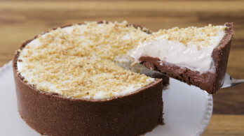 A cheesecake with a thick chocolate crust and a creamy white top layer adorned with crumbled cookie bits. A slice is being lifted from the cheesecake, showing a rich chocolate filling beneath the top layer. The cake is on a white plate with a wooden background.