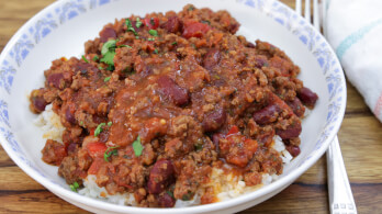 A white bowl filled with chili con carne, consisting of ground meat, kidney beans, and tomatoes, is placed on a wooden table. The chili is garnished with chopped parsley and served over a bed of white rice. A fork and a cloth napkin are positioned beside the bowl.