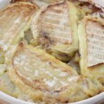 A round, white dish filled with a freshly baked gratin featuring golden, melted cheese slices on top of thinly sliced potatoes. The dish has browned edges, suggesting it has been oven-baked to a crispy finish. The background is a wooden table.