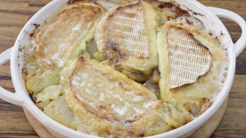 A round, white dish filled with a freshly baked gratin featuring golden, melted cheese slices on top of thinly sliced potatoes. The dish has browned edges, suggesting it has been oven-baked to a crispy finish. The background is a wooden table.