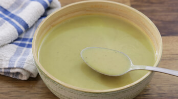 A ceramic bowl filled with a creamy green soup, with a metal spoon resting on the bowl's edge containing a portion of the soup. A folded white and blue checkered cloth is placed to the left of the bowl, all set on a wooden surface.