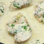 Chicken breasts cooking in a creamy sauce with herbs in a skillet. The sauce looks rich and smooth, generously coating the chicken, which is garnished with chopped parsley.