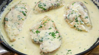 Chicken breasts cooking in a creamy sauce with herbs in a skillet. The sauce looks rich and smooth, generously coating the chicken, which is garnished with chopped parsley.