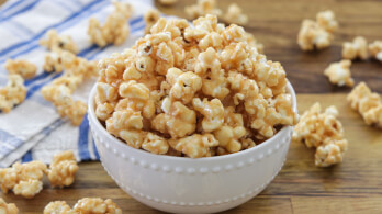 A white bowl filled with caramel popcorn sits on a wooden surface. Additional pieces of popcorn are scattered around the bowl. A blue and white checkered cloth is partially visible in the background.