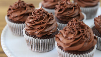 Close-up of several chocolate cupcakes with rich chocolate frosting swirled on top, placed in white paper liners. The cupcakes are arranged on a white plate, with a blurred background of a wooden surface.