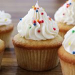 A group of vanilla cupcakes topped with swirls of white frosting and colorful sprinkles is arranged on a wooden surface. The cupcakes have light golden-brown exteriors and the frosting is piped in a decorative manner.