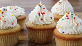 A group of vanilla cupcakes topped with swirls of white frosting and colorful sprinkles is arranged on a wooden surface. The cupcakes have light golden-brown exteriors and the frosting is piped in a decorative manner.