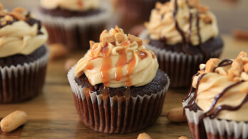 Seven chocolate cupcakes topped with creamy caramel frosting and drizzled with caramel and chocolate syrup. Each cupcake is garnished with chopped nuts, and they are arranged on a wooden surface with some whole peanuts scattered around.