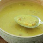 A bowl of yellow lentil soup with chunks of vegetables, shown with a spoonful lifted above the bowl. There is a blue and white checkered cloth in the background. The soup looks thick and hearty.