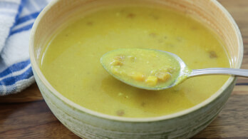 A bowl of yellow lentil soup with chunks of vegetables, shown with a spoonful lifted above the bowl. There is a blue and white checkered cloth in the background. The soup looks thick and hearty.