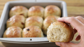 A person is holding a soft, light brown dinner roll with a bite taken out of it. In the background, a baking pan filled with similar rolls is visible, resting on a wooden surface. The rolls appear freshly baked and slightly golden.