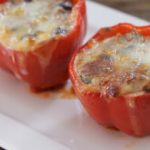 Three stuffed red bell peppers are placed in a row on a rectangular white plate. The peppers are filled to the brim with a mixture of rice, vegetables, and melted cheese that has turned golden brown on top. The dish appears freshly baked.