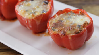 Three stuffed red bell peppers are placed in a row on a rectangular white plate. The peppers are filled to the brim with a mixture of rice, vegetables, and melted cheese that has turned golden brown on top. The dish appears freshly baked.