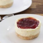 A small cheesecake with a crumbly crust and a red fruit topping sits on a white plate. In the background, another similar cheesecake is partially visible on another plate, with two spoons placed beside it on a wooden surface.