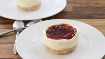 A small cheesecake with a crumbly crust and a red fruit topping sits on a white plate. In the background, another similar cheesecake is partially visible on another plate, with two spoons placed beside it on a wooden surface.