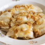 A white decorative plate filled with pierogies topped with caramelized onions and a light sauce. The plate has delicate floral patterns and a gold-trimmed edge. The pierogies appear soft, stuffed, and evenly coated with the golden-brown onions.