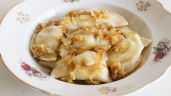 A white decorative plate filled with pierogies topped with caramelized onions and a light sauce. The plate has delicate floral patterns and a gold-trimmed edge. The pierogies appear soft, stuffed, and evenly coated with the golden-brown onions.