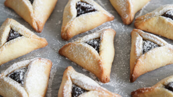 A close-up of several Hamantaschen cookies on a baking sheet. These triangular pastries are filled with a dark jam and dusted with powdered sugar. The cookies are arranged neatly in rows.