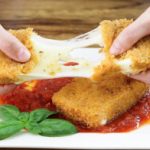 Hands pulling apart a piece of breaded fried cheese, with melted cheese stretching between the pieces. The cheese is on a white plate with tomato sauce and a basil garnish underneath. The background is a wooden surface.