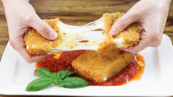 Hands pulling apart a piece of breaded fried cheese, with melted cheese stretching between the pieces. The cheese is on a white plate with tomato sauce and a basil garnish underneath. The background is a wooden surface.